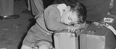 Black and white archival image of a young immigrant boy asleep on a pair of suitcases at Pier 21